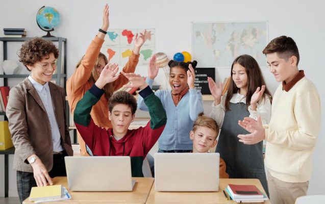 Group of young students at school sitting and standing in front of two laptop computers celebrating.