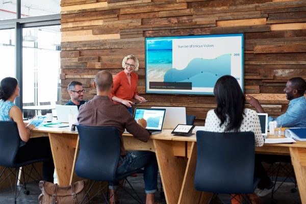 A woman standing next to a display, speaking to five people with computers or tablets in a huddle space.